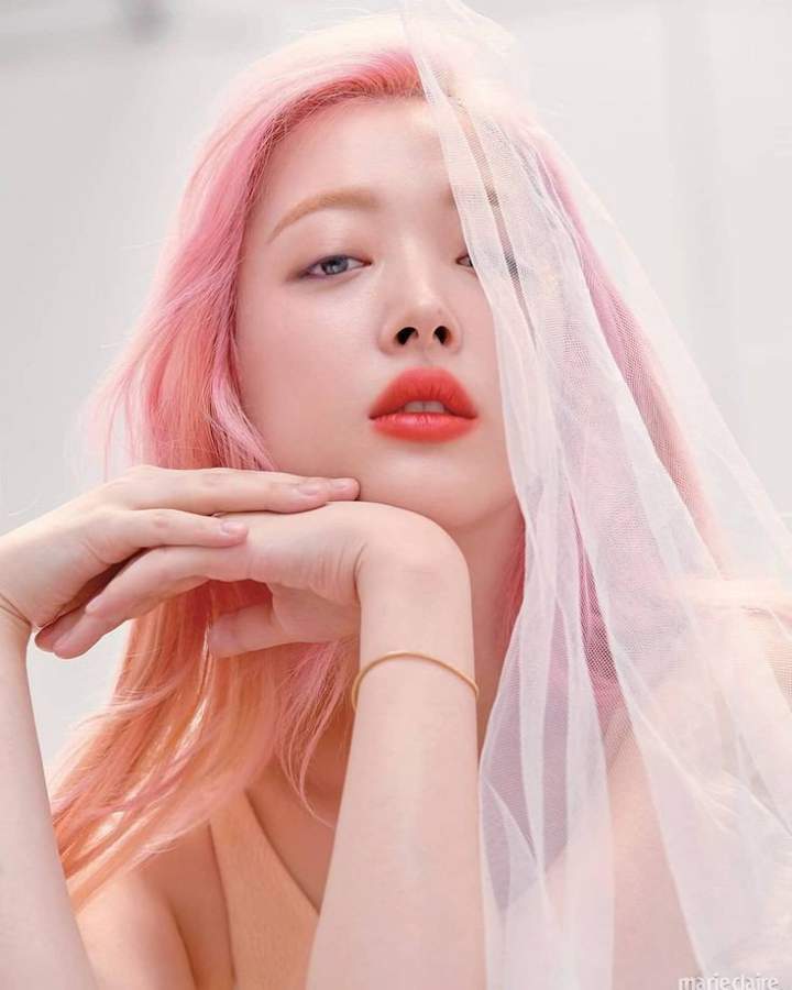 image of kpop star sulli. she has pink/peach hair and is looking at the front of the image with her head tilted upward. her hands are under the right side of her chin. the image is lightly colored.