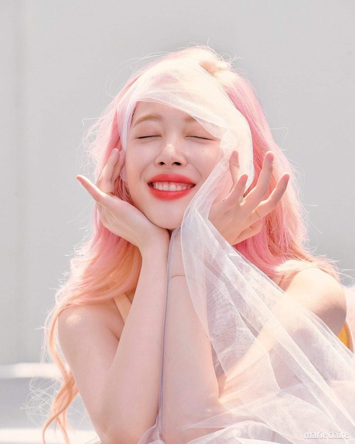 image of kpop star sulli. she has pink/peach hair and is turned to the left of the image. her eyes are closed and her hands are on the sides of her face, she is smiling brightly. the image is lightly colored.